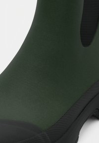 Zielony gumowy but o matowym wykończeniu, z teksturowaną czarną podeszwą i otworami wentylacyjnymi po bokach. Posiada okrągły czubek i bezszwowy design cholewki.
