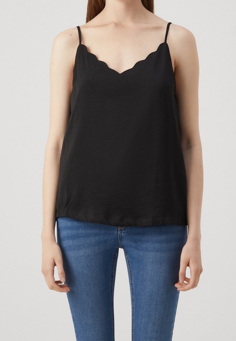 Femme portant un haut noir sans manches avec un col festonné et un jean bleu, debout devant un fond clair uni.