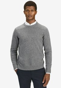 Grijze wollen trui met een ronde hals en ribgebreide manchetten en hem, gedragen over een witte blouse met kraag. Model staat tegen een witte achtergrond.