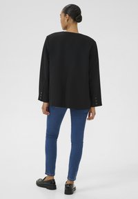 Čierny oversized blejzer so striedmym strihom, minimálnymi švami a gombíkovými manžetami, v kombinácii s modrými skinny džínami a čiernymi mokasínami.