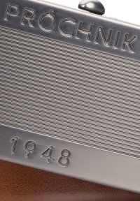 Metaliczna kopertówka z wytłoczonym napisem "PRÓCHNIK" i poziomymi liniami; zawiera wygrawerowany rok "1948", osadzony na brązowym skórzanym tle o fakturze.