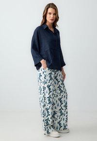 Chemise en lin bleu marine avec une fermeture à boutons à l'avant et des manches larges, assortie à un pantalon blanc à motif floral bleu. Chaussures décontractées incluses.