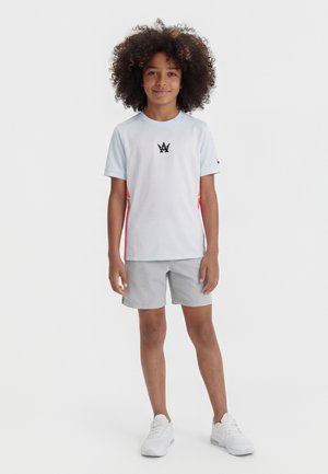 Παιδί με σγουρά μαλλιά στέκεται μπροστά, φορώντας λευκό κοντομάνικο μπλουζάκι με λογότυπο, γκρι σορτς και λευκά αθλητικά παπούτσια σε λευκό φόντο.