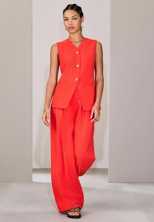 Kvinde med flettet hår, iført rød vest uden ærmer med guldknapper og matchende bukser med brede ben, går i et rum med neutrale toner.