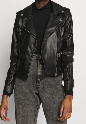 Personne portant une veste de motard en cuir noir avec des fermetures éclair argentées sur un haut noir et un jean gris taille haute.