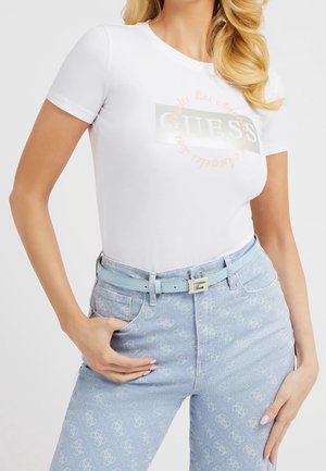 White fitted t-shirt featuring a glossy "GUESS" logo and pastel accents, paired with light blue jeans that have a logo pattern and a matching belt.