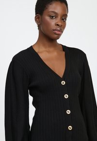 Schwarzer Cardigan mit V-Ausschnitt, gerippter Textur und langen Ärmeln. Verfügt über fünf goldene Knöpfe auf der Vorderseite und ein gestricktes Muster an den Ärmeln.