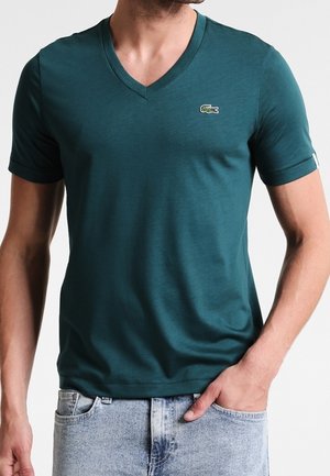 Mann trägt ein dunkel-türkisfarbenes V-Ausschnitt Kurzarm-T-Shirt mit einem kleinen Krokodillogo auf der linken Brust, kombiniert mit hellblauen Jeans.