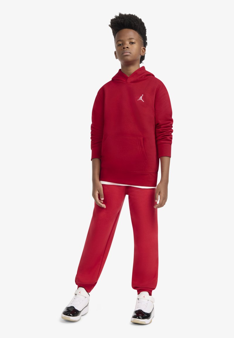 Rdeča jopa s kapuco z žepom spredaj in belim logotipom, združena z ujemajočimi se trenirkami. Obutev ima črno-bele poudarke.