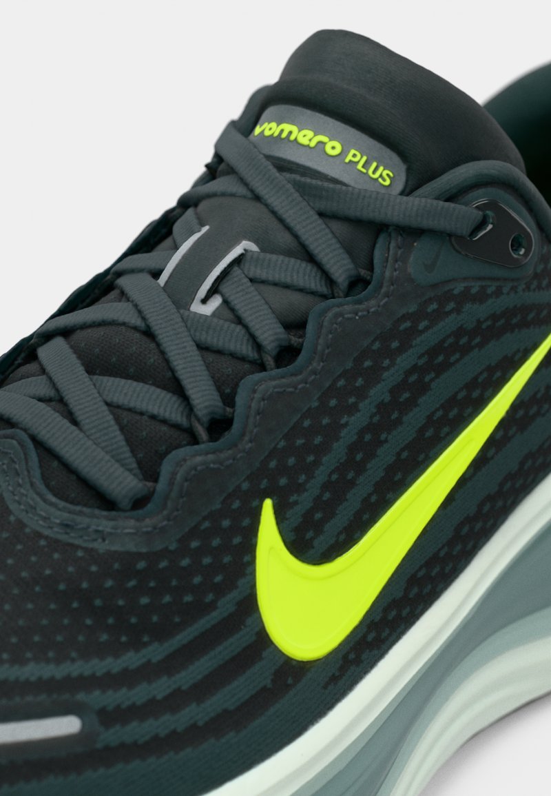 Primer plano de unas zapatillas Nike Vomero Plus en negro y gris con un swoosh en amarillo neón y un logo a juego en la lengüeta acolchada.