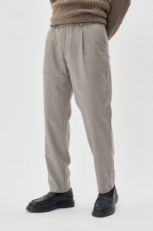 Pantalon gris clair avec un tissage texturé, un devant plissé et des poches latérales, associé à des mocassins noirs sur un fond uni.