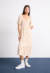 Lys beige blomstrete kjole med korte flagrende ermer, V-hals, og et lagdelt skjørt, sammen med hvite sneakers og ankelsokker.
