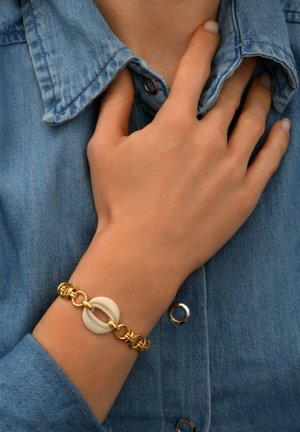 Bracelet doré avec un accent circulaire blanc, ayant des maillons interconnectés et une surface texturée, porté sur une chemise en denim.
