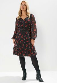 Robe floral noire avec de longues manches transparentes, décolleté en V, détail de ceinture cloutée et ourlet à volants superposés. Portée avec des bottines noires.