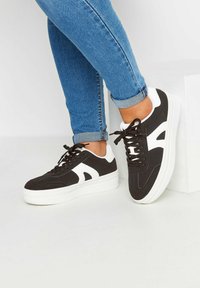 Svarta och vita sneakers med överdel i mocka och läder, snörning fram och tjock vit plattformssula; sidologotyp synlig.