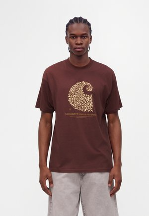 Νέος άνδρας με πλεξούδες, φοράει ένα καφέ t-shirt Carhartt με αφηρημένο κίτρινο κυματιστό σχέδιο και ανοιχτό γκρι παντελόνια, στέκεται μπροστά σε λευκό φόντο.