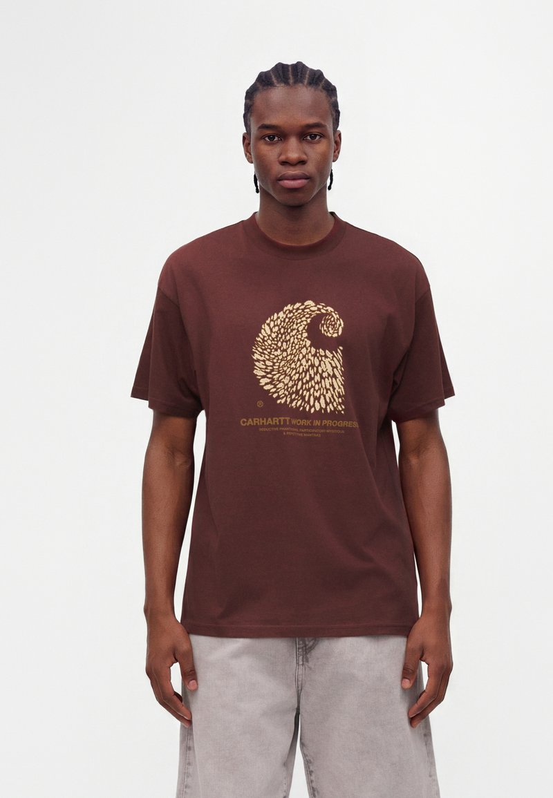 Νέος άνδρας με πλεξούδες, φοράει ένα καφέ t-shirt Carhartt με αφηρημένο κίτρινο κυματιστό σχέδιο και ανοιχτό γκρι παντελόνια, στέκεται μπροστά σε λευκό φόντο.