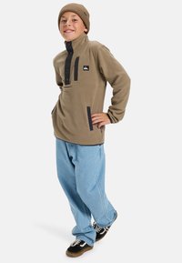 Pull en polaire marron avec un col haut, patte de boutonnage noire, poche avant et accents contrastants noirs, associé à un jean bleu clair.