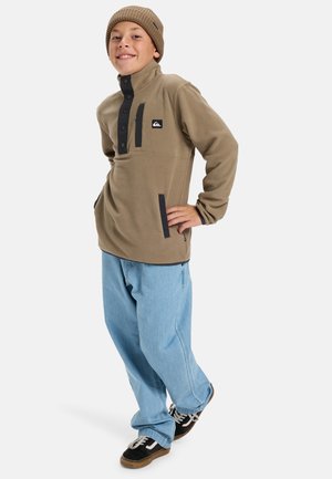 Sudadera de forro polar marrón con cuello alto, tapeta de botones negra, bolsillo frontal y detalles en negro que contrastan, combinada con jeans azules claros.