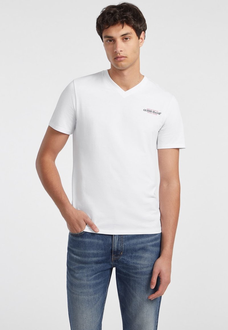 Biely tričko s v-výstrihom vyrobené z bavlny s krátkymi rukávmi, s malým červeným logom "GUESS JEANS" na ľavej hrudi. Nosené s modrými džínsami.