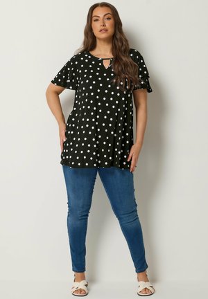 Vrouw met lang bruin haar die een zwarte tuniek met korte mouwen en polkadots draagt, blauwe skinny jeans en witte sandalen, staand tegen een effen achtergrond.