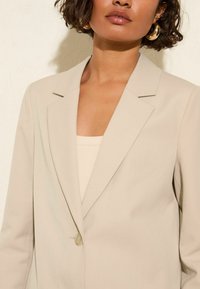 Béžový dobře střižený blazer s jedním knoflíkem, klopovými límečky a hladkou texturou. Nosený přes světlý tílko, zdůrazňující minimalistický design.
