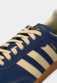 Μπλε σουέντ αθλητικό παπούτσι Adidas Samba με μπεζ κορδόνια, τρεις μπεζ ρίγες στο πλάι και σόλα από καουτσούκ gum ορατή.