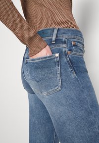 Personne portant un jean en denim bleu avec un haut à manches longues marron à motifs, main placée dans la poche arrière du jean.