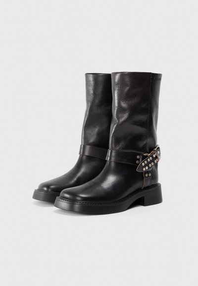 Bottes en cuir noir montant jusqu'au genou avec une finition texturée, présentant une sangle brun foncé ornée de clous en argent au niveau de la cheville. Design à semelle épaisse.