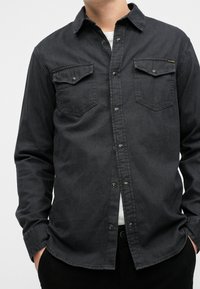 Camicia di denim nero con due tasche sul petto, bottoni a pressione, colletto appuntito e maniche lunghe. Il tessuto appare leggermente strutturato e resistente.