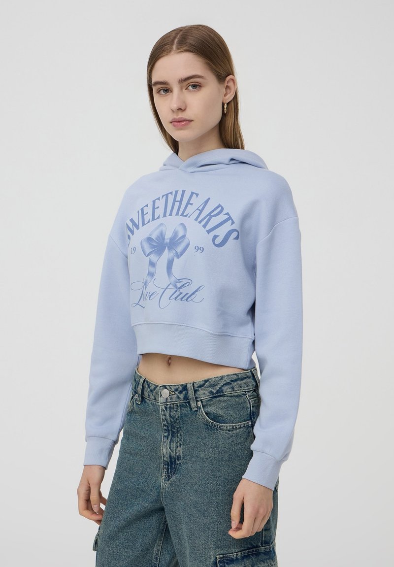 Felpa cropped di un blu chiaro con cappuccio e lacci, caratterizzata da una stampa grafica di fiocchi e testo in blu scuro. Tessuto morbido, vestibilità comoda.