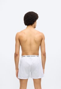 Άσπρα καρό μποξεράκια με μαύρη ζώνη μέσης με λογότυπο, εμφανιζόμενα από πίσω σε ένα άτομο με σγουρά μαλλιά, γυμνούς ώμους και χωρίς πουκάμισο.