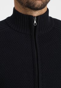 Tmavě pletený svetr s vysokým žebrovaným límcem, plným zipem vpředu a strukturovanou tkaninou, v tmavě modré barvě.