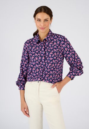 Marineblouse met een bladerenpatroon in roze en wit, met een knoopsluiting bij de kraag, lange mouwen en opgerolde manchetten, gecombineerd met beige broek.