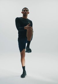 Schwarzes Langarmshirt, blaue Shorts und schwarze Sportschuhe. Die Person dehnt sich mit einem erhobenen Knie. Sauberer, neutraler Hintergrund.
