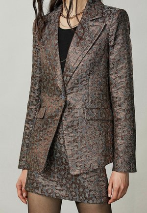 Žena v metalickém blejzru se vzorem leopardí kůže a odpovídající sukni přes černý top a průhledné černé punčochy, stojící před jednobarevným pozadím.