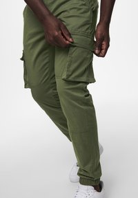 Groene cargo broek met zijzakken, taps toelopend ontwerp, elastische boorden en duurzame stof. Model draagt witte sneakers.