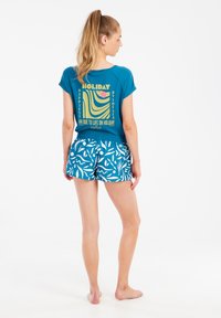 Žalias trumpomis rankovėmis marškinėliai su grafiniu spaudiniu ant nugaros, derinami su raštuotais mėlynais šortais, turinčiais baltus abstrakčius raštus.