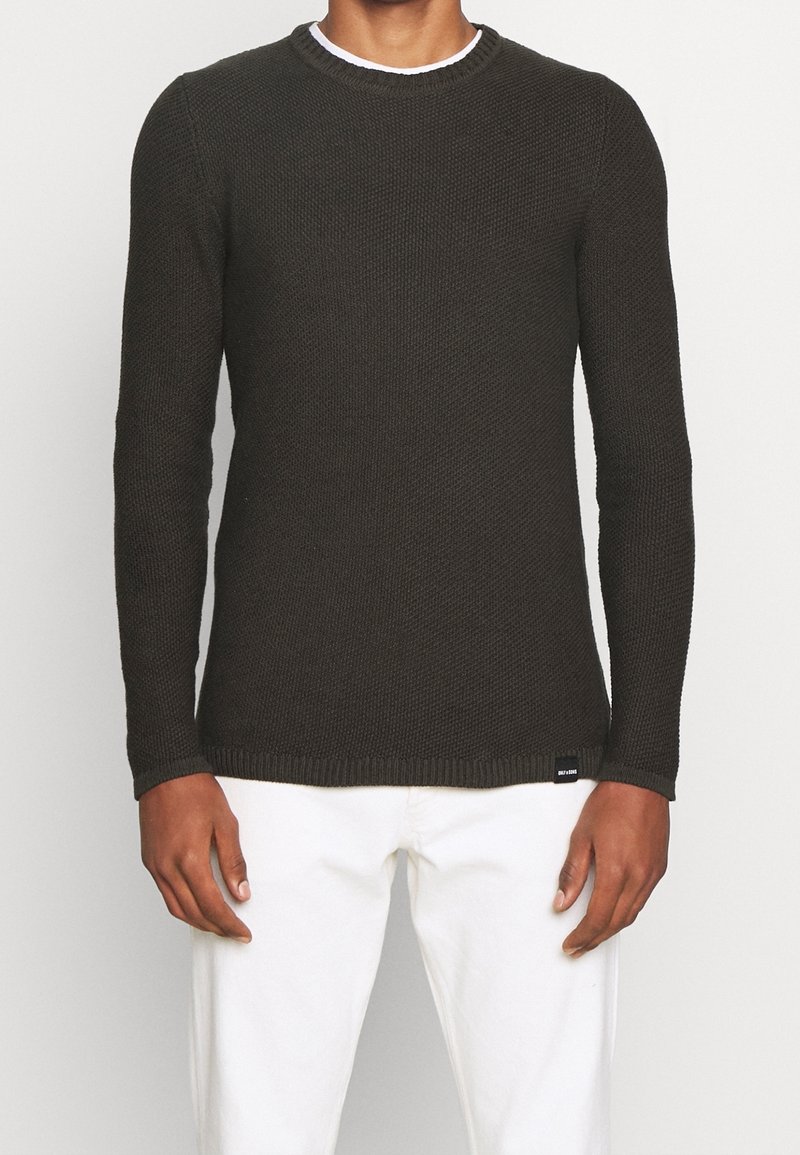 Homme portant un pull en maille texturée vert foncé et un pantalon blanc, debout devant un fond clair uni.