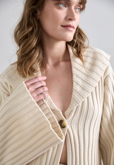 Beige ribbed cardigan with wide sleeves and large button closure. Features a deep V-neckline and textured pattern throughout the fabric.