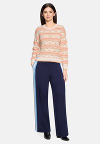Pull en maille rayé crème avec des vagues corail et beige ; associé à un pantalon large bleu marine avec des bandes latérales bleu clair.