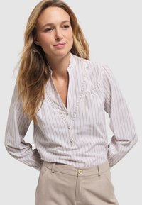 Biała koszula z cienkimi pionowymi paskami w kolorze beżowym, z teksturowanym koronkowym wykończeniem wzdłuż klatki piersiowej i falbaniastym kołnierzykiem. Połączona z jasnymi spodniami.