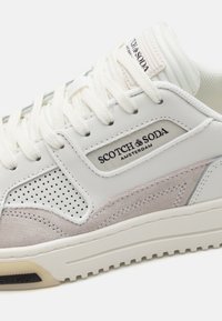 Hvide lædersneakers med grå ruskindsdetalje, perforerede sidepaneler og polstret krave. Brandlogoet vises på siden.