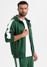 Homme portant un survêtement vert et blanc avec une veste à capuche et un T-shirt blanc, posant devant un fond clair uni.