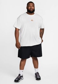 White cotton t-shirt with orange logo, paired with black shorts. Worn with gray and black sneakers, white socks, and a short, trimmed beard.
