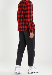 Persoon in een rood en zwart geruit overhemd, zwarte relaxed-fit jeans, witte sokken en witte Nike sneakers die zijwaarts staat.