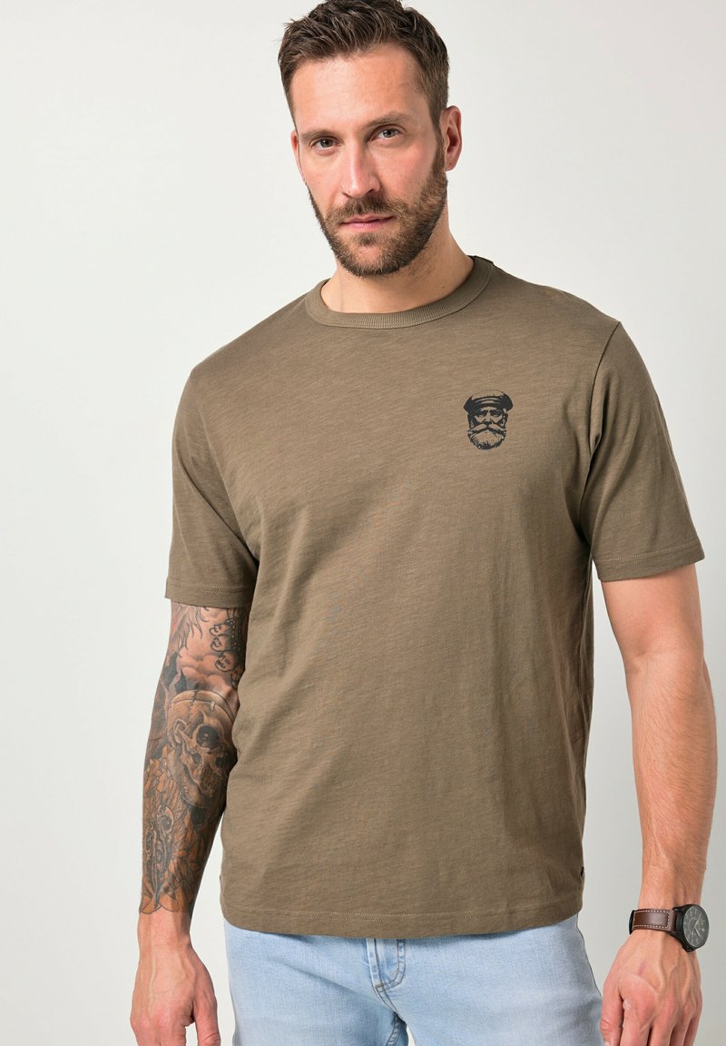 Mand med skæg og tatoveret venstre arm iført brun t-shirt med sort sømandssymbol og lyseblå jeans, stående mod en ensfarvet baggrund.