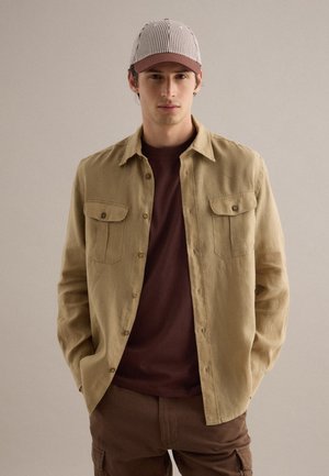 Mann trägt beige Hemd über dunkelbraunem T-Shirt, braune Cargohose und gestreifte Mütze, steht mit Händen in den Taschen.