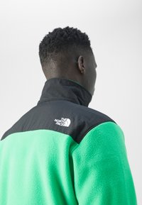 Vyras su trumpais garbanotais plaukais, vilkintis žalią ir juodą vilnos striukę su "The North Face" logotipu, atsigręžęs nuo vientiso fono.