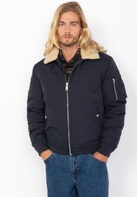 Chaqueta bomber de navy con un cuello de borrego beige, cierre frontal con cremallera, puños acanalados y un bolsillo lateral con cremallera, llevada sobre una camisa de cuadros.